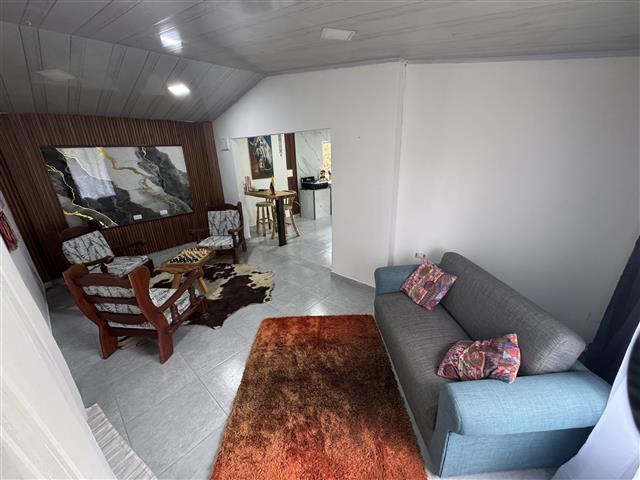 eco-hotel-gaia-15 - Turismo en el Eje Cafetero, Alquiler de Fincas en el Quindio Cozy living room with a gray-blue sofa and pink cushions, an orange rug, and mid-century chairs around a coffee table; doorway leads to another room with a rug.