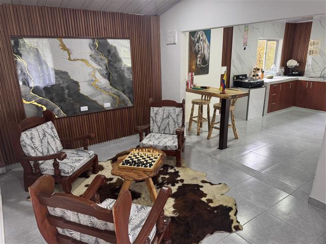 eco-hotel-gaia-16 - Turismo en el Eje Cafetero, Alquiler de Fincas en el Quindio Cozy seating area with four wooden armchairs around a small chessboard on a wooden table atop a cowhide rug; kitchen with bar stools in back.