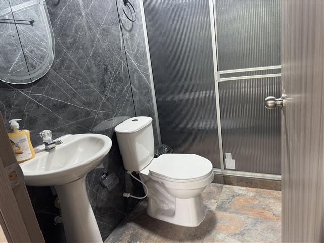 eco-hotel-gaia-18 - Turismo en el Eje Cafetero, Alquiler de Fincas en el Quindio Small bathroom with a white pedestal sink, toilet, and a frosted glass shower door; dark marble-patterned walls and a bottle of hand soap on the sink surface.