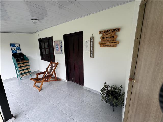 eco-hotel-gaia-21 - Turismo en el Eje Cafetero, Alquiler de Fincas en el Quindio Covered porch with a wooden rocking chair, a teal drawer cabinet, a dark door, welcome signs on the wall, a potted plant, and tiled flooring.